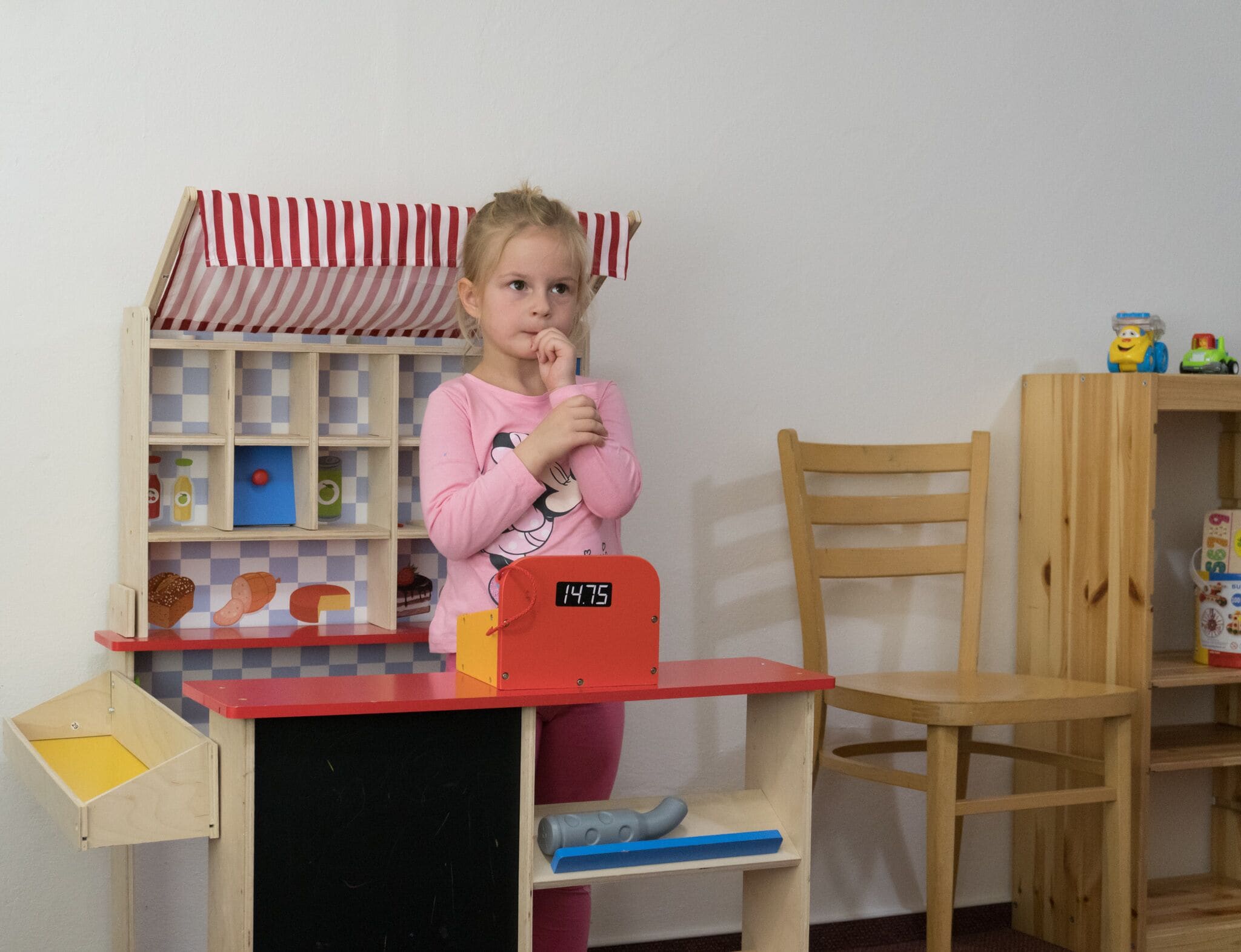 Niño en la caja de una tienda de juguetes, con un toldo blanco y rojo, una silla de madera y una estantería con varios artículos.