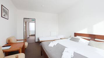 A hotel room with a large bed, a smaller bed, a desk, and a chair. There is a framed picture on the wall and a door leading to another room.