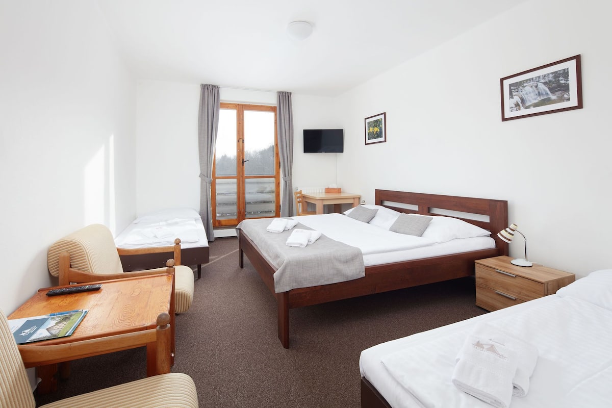 Quarto de hotel com cama, escrivaninha, cadeira, televisão e janela com cortinas.