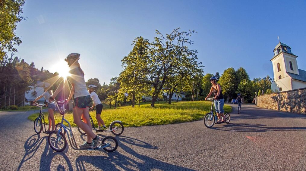 A group of people riding bicycles and unicycles on a paved path in a park with a church in the background.