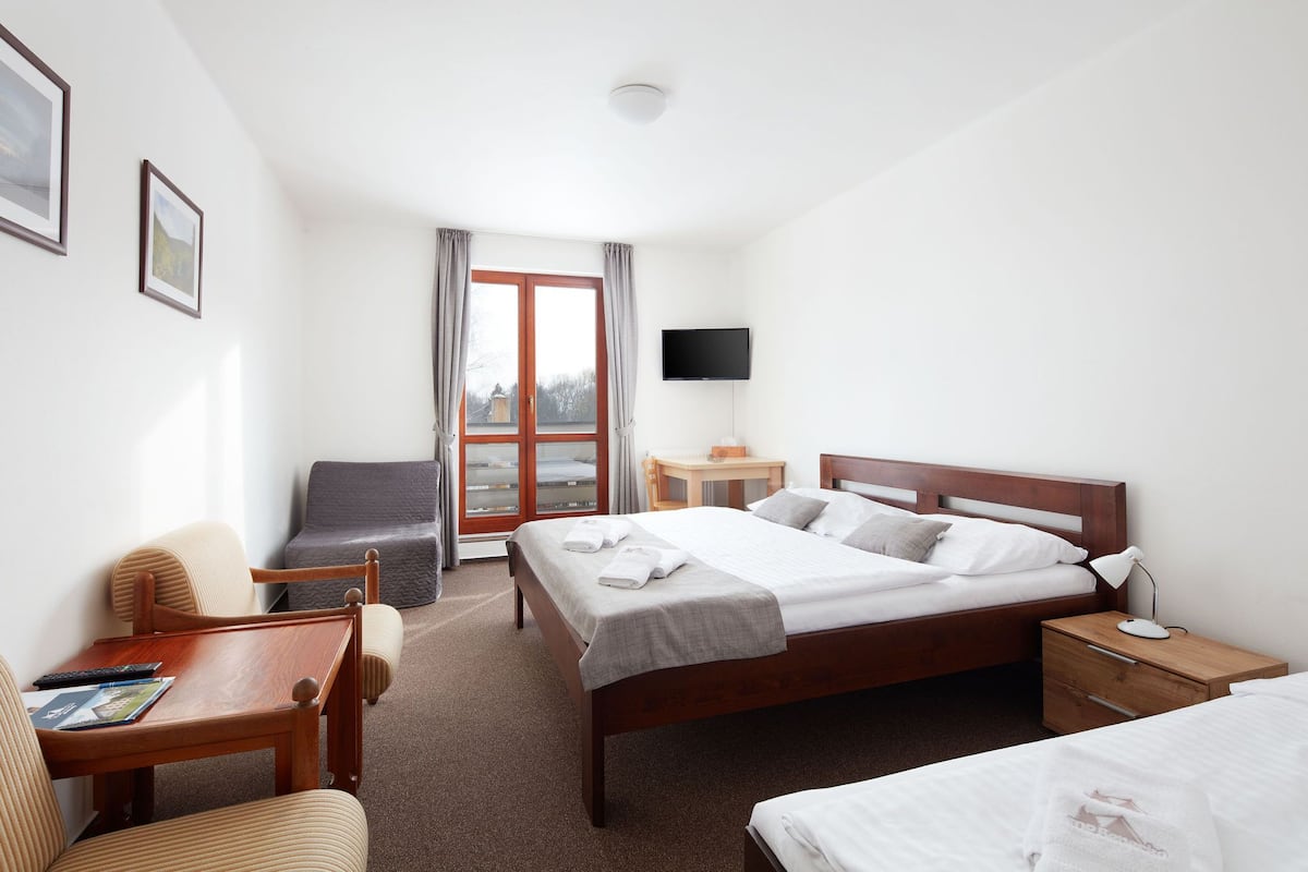 Quarto de hotel com cama, escrivaninha, cadeira, televisão e janela com cortinas.