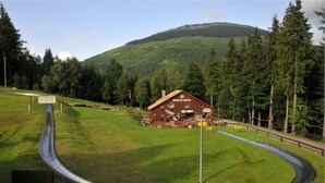 Een landelijke omgeving met een huis, genaamd 'Bobová Dřeň', omringd door een omheind gebied, een kronkelend pad en een berg op de achtergrond.