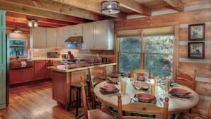 A wooden cabin interior with a kitchen and dining area, featuring wooden beams, a large window, and a dining table set for four. - Creek-front Cabin on Wooded Acres (Sugar Grove)