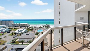 A high-rise building with a balcony overlooking a beach, pool, and adjacent buildings.