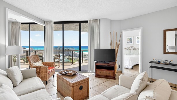 A modern living room with a large TV, wicker furniture, a wicker coffee table, and a view of the ocean.
