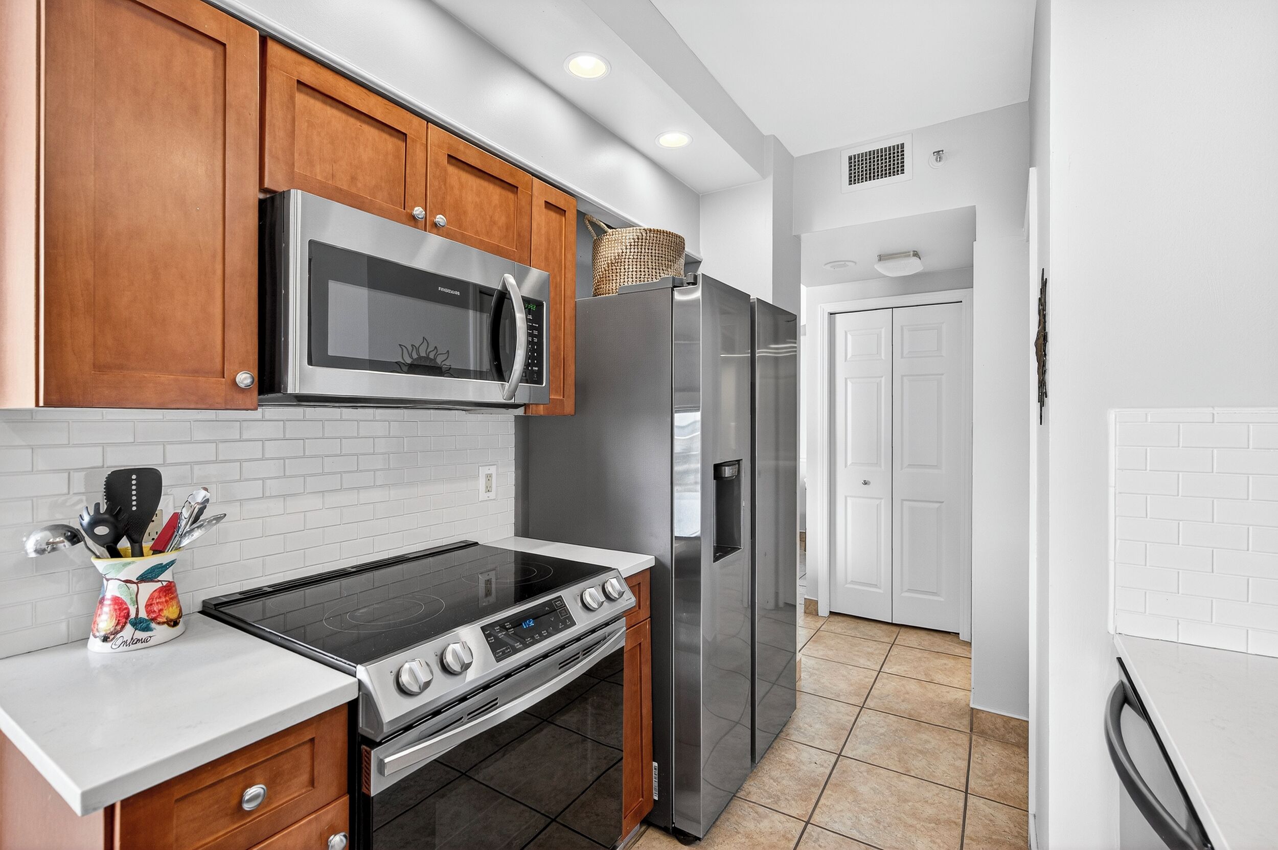 A kitchen with wooden cabinets, a stainless steel microwave, a refrigerator, a stove, and a white tiled backsplash.