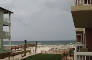 A beach view from a balcony with a wooden deck, a small pool, and a fenced area with palm trees. - HEATED  POOL  AND  HOT  TUB (Orange Beach)