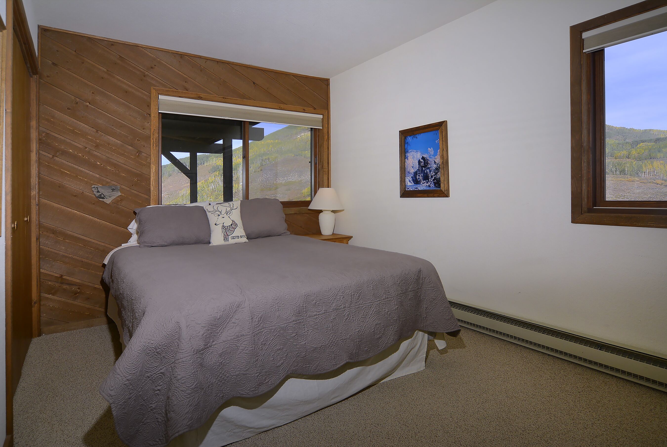 Un dormitorio con cama, mesita de noche, lámpara, un cuadro enmarco y ventana con vistas a árboles y colinas.