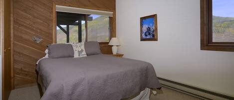 Un dormitorio con cama, mesita de noche, lámpara, un cuadro enmarco y ventana con vistas a árboles y colinas.
