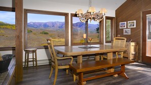 Un coin repas avec une table en bois, des chaises et un lustre. De grandes fenêtres offrent une vue sur les montagnes.