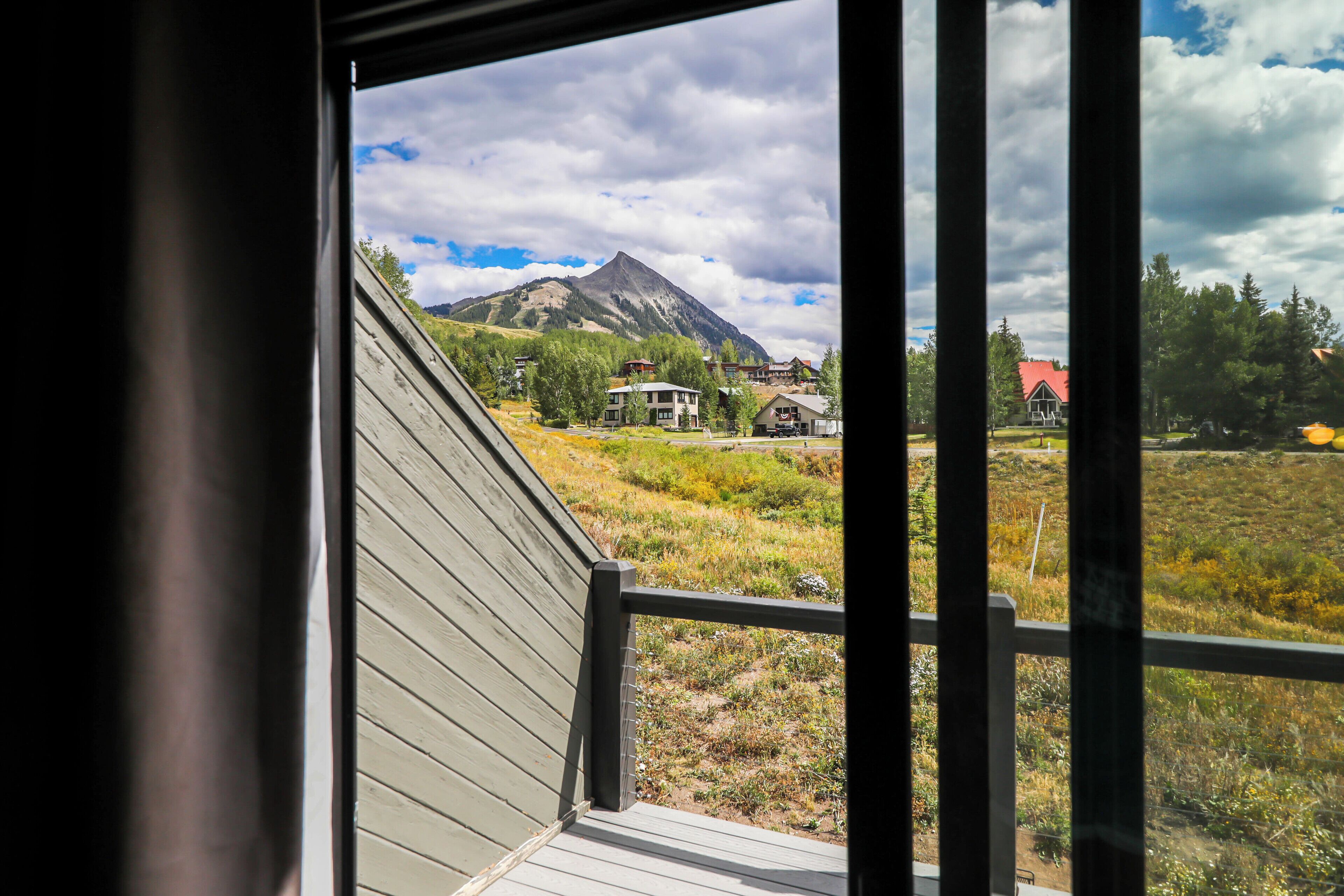 A view from a window with a wooden deck, overlooking a mountainous landscape with houses and a clear sky.