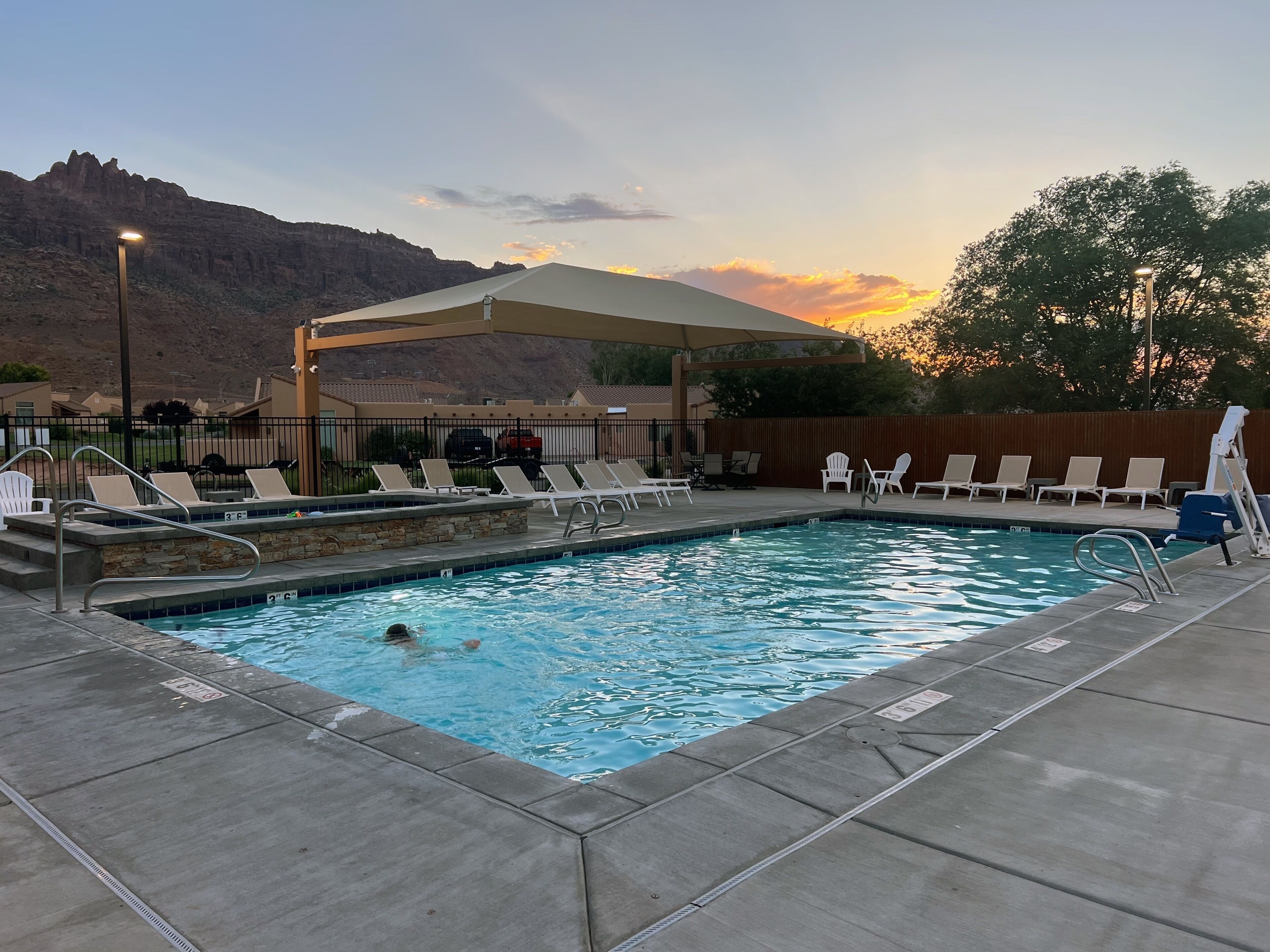 Piscina con sillas de descanso y una zona cubierta, rodeada por una valla y un paisaje montañoso.