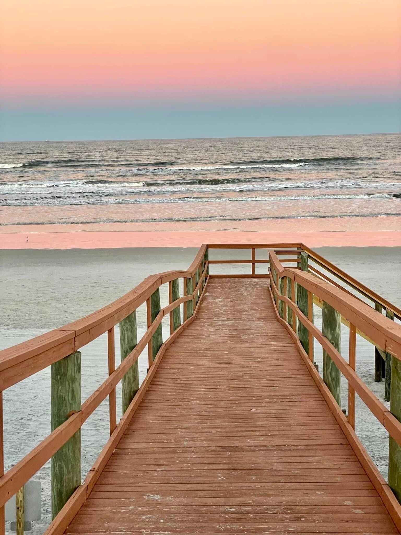 Un paseo marítimo de madera que conduce a una playa con el atardecer al fondo.