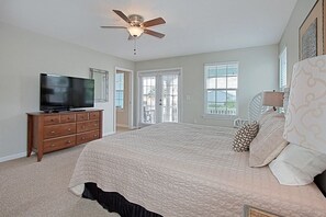 A bedroom with a bed, a TV on a wooden dresser, a ceiling fan, and a window with blinds.