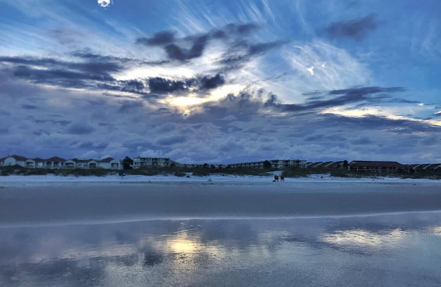 Une plage avec une surface d’eau qui reflète l’environnement, des bâtiments en arrière-plan et un ciel nuageux.