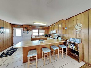 A kitchen with wooden cabinets, a refrigerator, a microwave, and a coffee maker. There are three bar stools and a bookshelf with books. - Both Sides Now | Ocean View + Walk to the Beach! (Topsail Beach)
