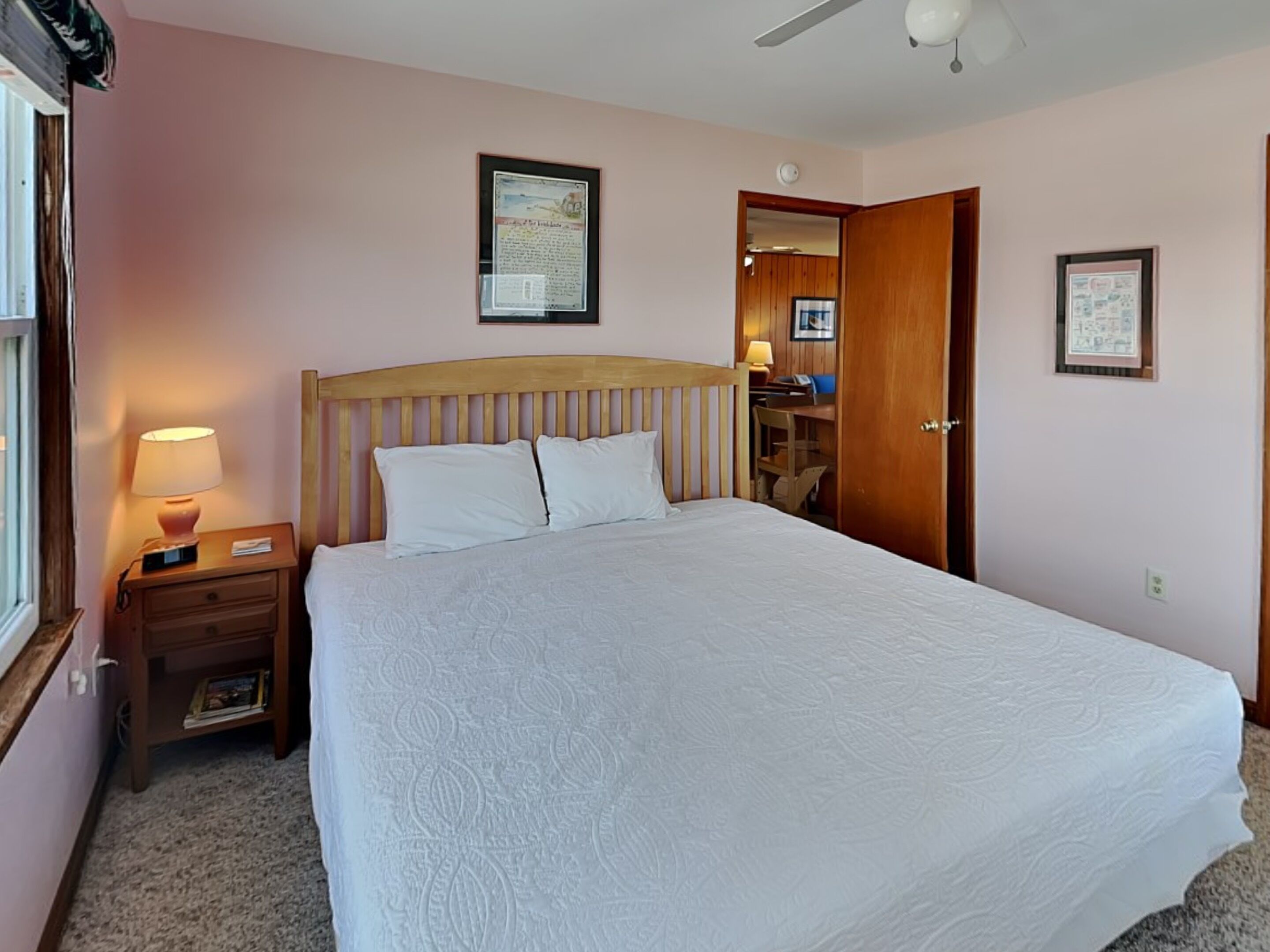 A bedroom with a bed, a nightstand, a lamp, a wooden headboard, a ceiling fan, and framed pictures on the walls.