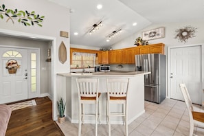A kitchen with wooden cabinets, a stainless steel refrigerator, a bar area with two white chairs, and a decorative wall piece.
