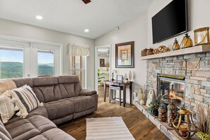 A living room with a sectional sofa, a fireplace, a flat-screen TV, and a view of the outdoors. - Best mountain view in TN! See 3 states from the deck! See the houses in valley? (Tallassee)