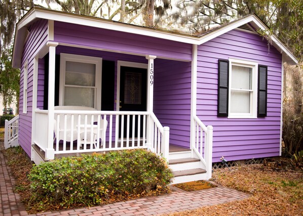 Une maison violette avec un porche blanc, un petit jardin devant et une boîte aux lettres.