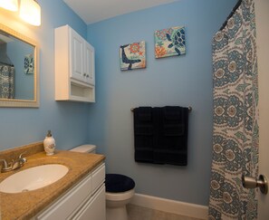 Une salle de bain équipée d’un lavabo, de toilettes et d’un porte-serviettes. On y trouve deux œuvres d’art accrochées au mur et un rideau de douche à motifs.