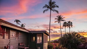 Une maison avec une véranda, un banc et une vue sur des palmiers se découpant sur un ciel au coucher du soleil.
