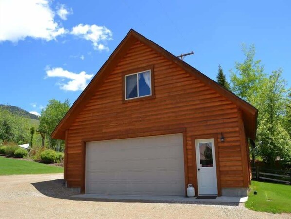 A wooden cabin with a garage, surrounded by greenery and a clear sky. - Cozy Retreat | 18 MI to YNP | Lake Views (Island Park)