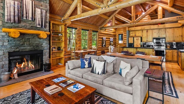 A log cabin interior with a fireplace, a sofa, a coffee table, and a kitchen area. - RMR: Granite Ridge Cabin 7586 in Teton Village (Teton Village)