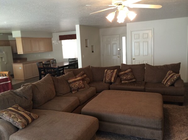 A living room with a sectional sofa, ottoman, and a dining area with a table and chairs. - Las Palmas resort ground floor 3 bedroom condo large couch seating hottubs pools (St George)