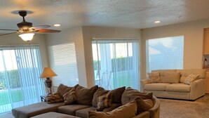 A living room with a large sectional sofa, a smaller sofa, and a coffee table. There is a ceiling fan, a lamp, and a window with blinds. - Las Palmas resort ground floor 3 bedroom condo large couch seating hottubs pools (St George)