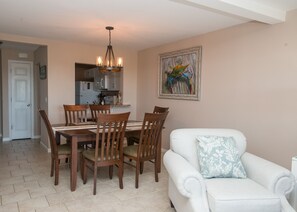 A dining area with a wooden table and chairs, a chandelier, a kitchen in the background, and a painting on the wall.