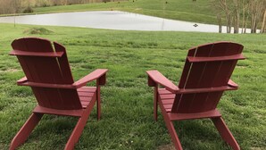 Two Adirondack chairs facing a pond with a view of a green field and hills in the background.