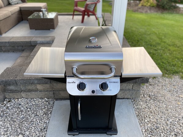Un barbecue à gaz Char-Broil doté d’un panneau de commande noir et d’un couvercle en acier inoxydable, installé sur une terrasse en béton bordée de pierres.