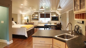A bedroom with a bed, bedside table, and a window. There is a kitchenette with a sink, stove, and cabinets.