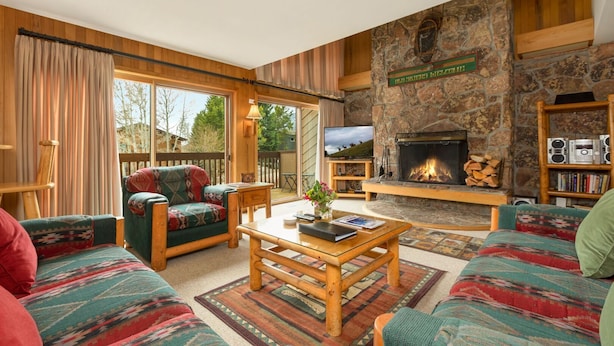 A cozy living room with a stone fireplace, wooden furniture, and a large window overlooking a deck.