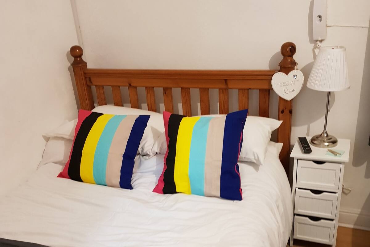 A bed with a wooden headboard, white bedding, and striped pillows. There is a bedside table with a lamp and a remote control.