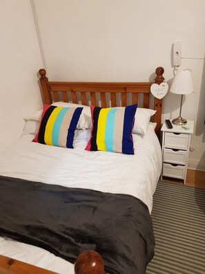 A bed with a wooden headboard, white bedding, and striped pillows. There is a bedside table with a lamp and a remote control. - Quality Accommodation (Leeds)