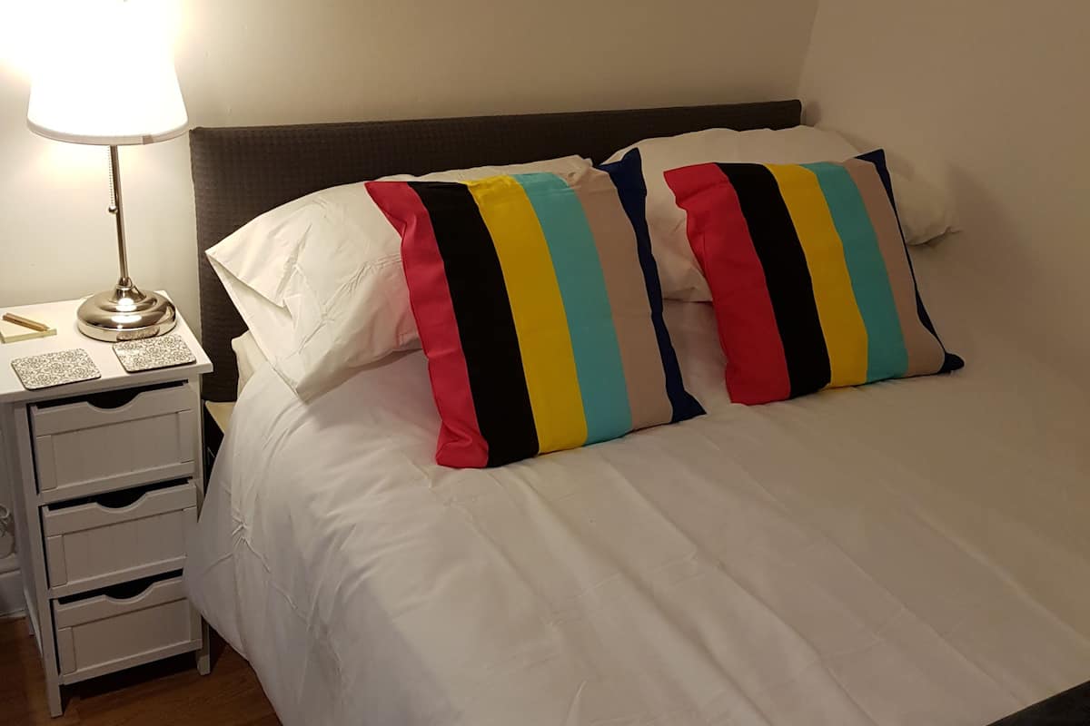 A neatly made bed with white bedding and colorful striped pillows, a bedside table with a lamp, and a small drawer unit.