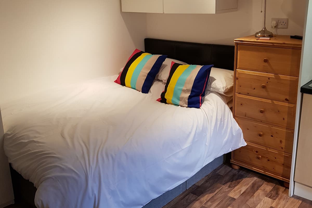 A neatly made bed with a white duvet, a wooden dresser with drawers, a lamp on the dresser, and a small rug on the floor.