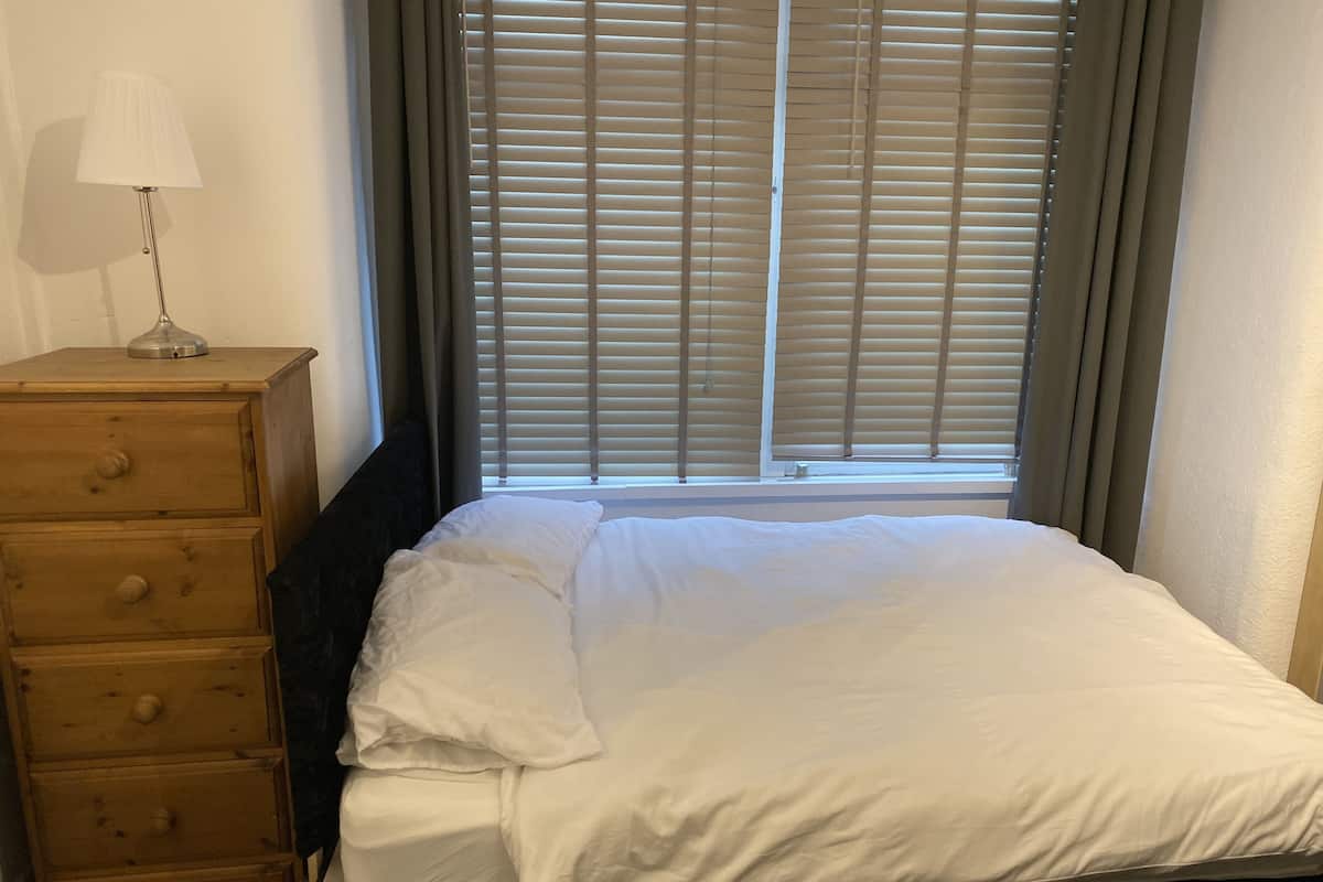 A neatly made bed with white bedding, a wooden dresser with drawers, and a window with blinds.