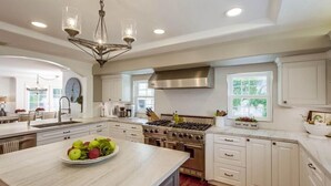 Una cocina amplia con armarios blancos, una isla grande con fruteros, electrodomésticos de acero inoxidable y una lámpara moderna.