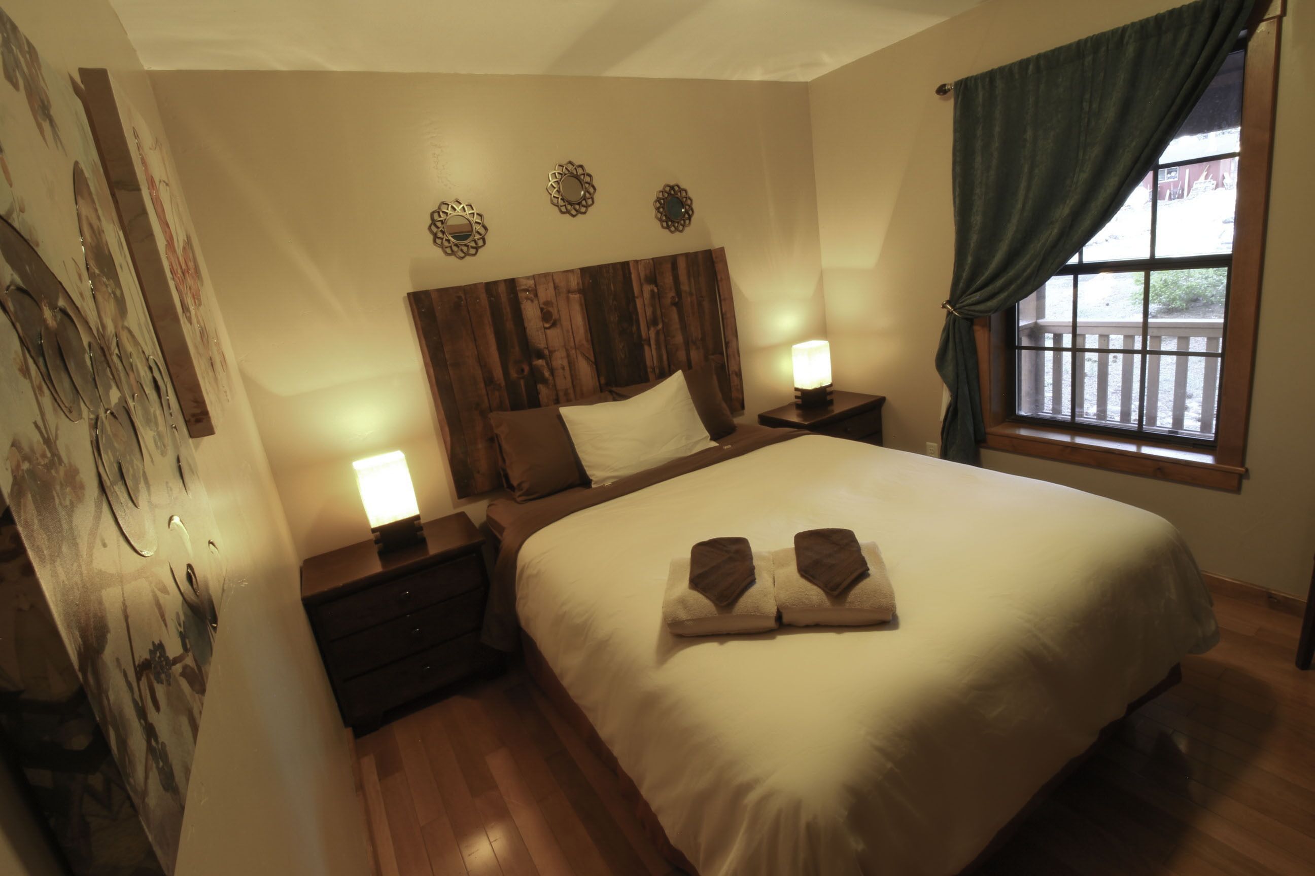 A bedroom with a wooden headboard, a large bed with two pillows, two bedside lamps, a window with a view, and a wall with decorative plates.