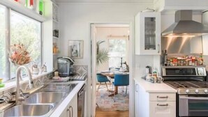A modern kitchen with a stainless steel sink, a stove, and a countertop with various items. There is a window with curtains and a door leading to another room. - Under the Oaks Santa Barbara (Santa Barbara)