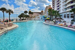 Una gran piscina al aire libre con sillones de descanso y sombrillas, rodeada de palmeras y un edificio de hotel con balcones.