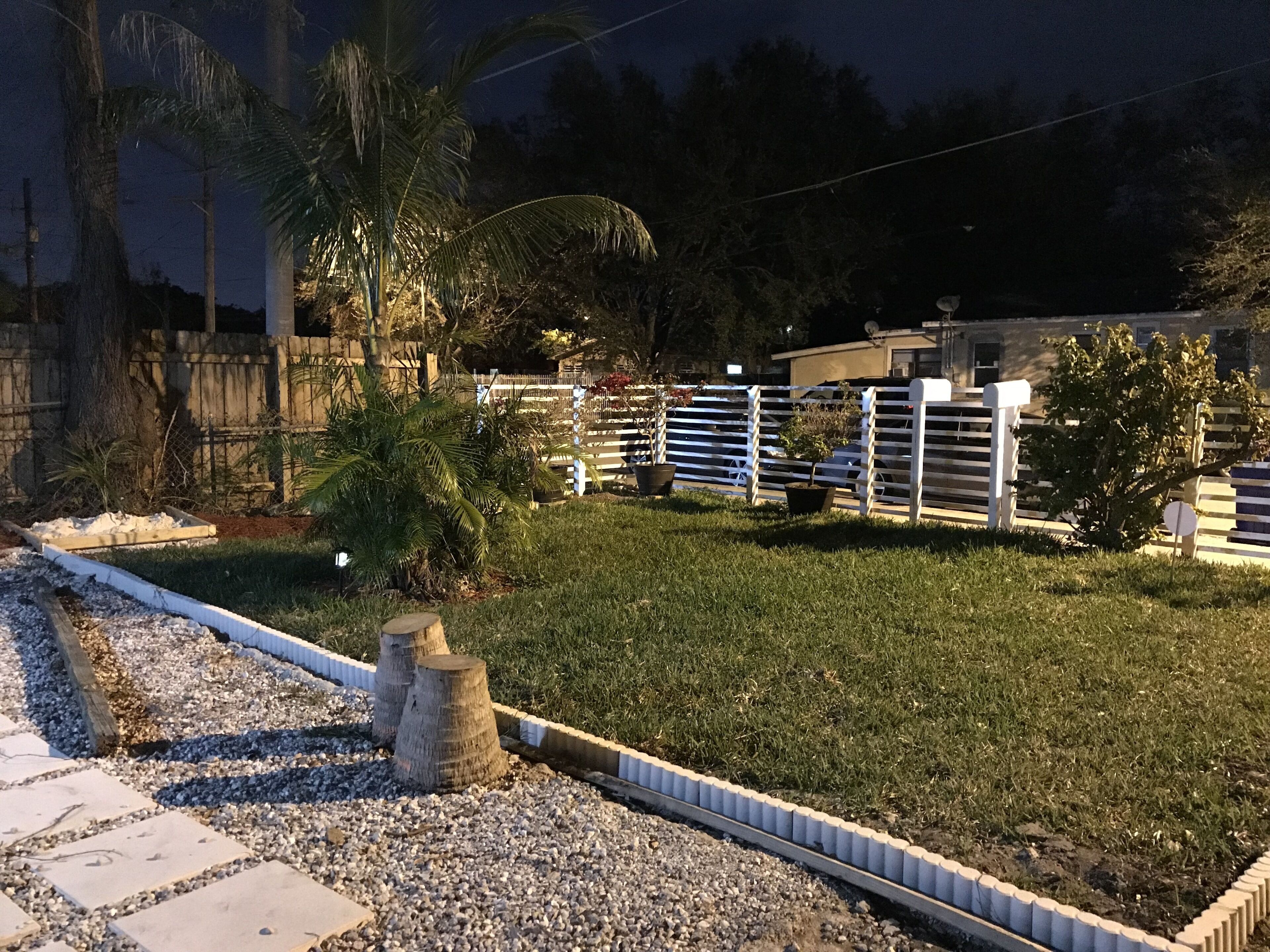 Un jardin bien entretenu, avec un chemin de gravier, un palmier et une clôture blanche.