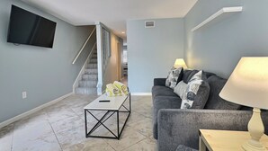 A living room with a grey sofa, a coffee table, a TV mounted on the wall, and a staircase leading to another room. - Walk to the #1 Beach in America, Offers Heated community Pool, Free Wi-Fi (Siesta Key)