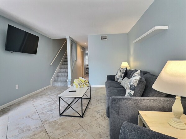 A living room with a grey sofa, a coffee table, a TV mounted on the wall, and a staircase leading to another room.