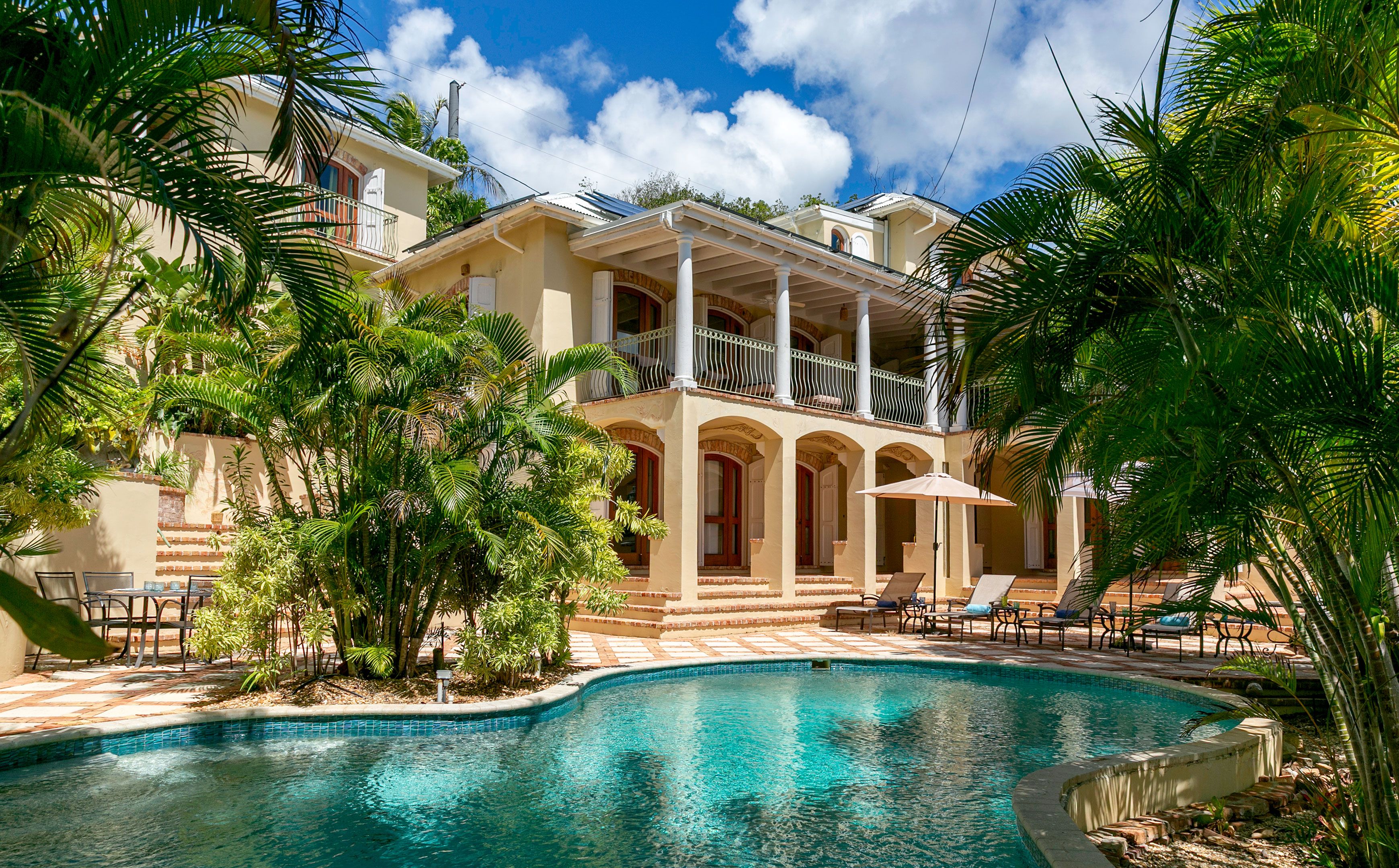 A poolside view of a hotel with a pool, lounge chairs, and tropical vegetation.