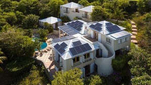 A modern house with multiple solar panels on the roof, surrounded by lush greenery and a swimming pool.