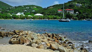 A beach with a rocky foreground, clear blue water, and houses on a hillside in the background.